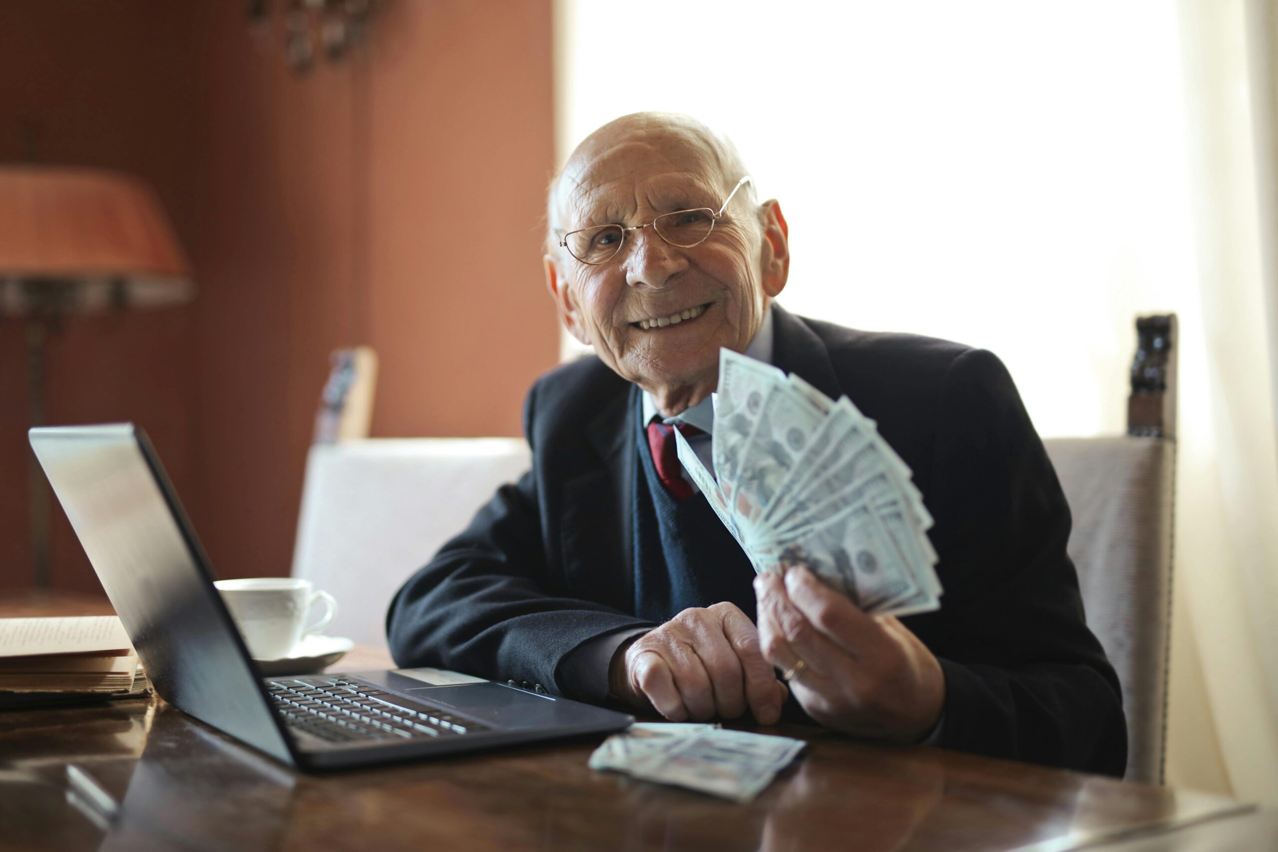 GlÃ¼cklicher Ã¤lterer GeschÃ¤ftsmann, Der Geld In Der Hand HÃ¤lt, WÃ¤hrend Er Am Laptop Am Tisch Arbeitet